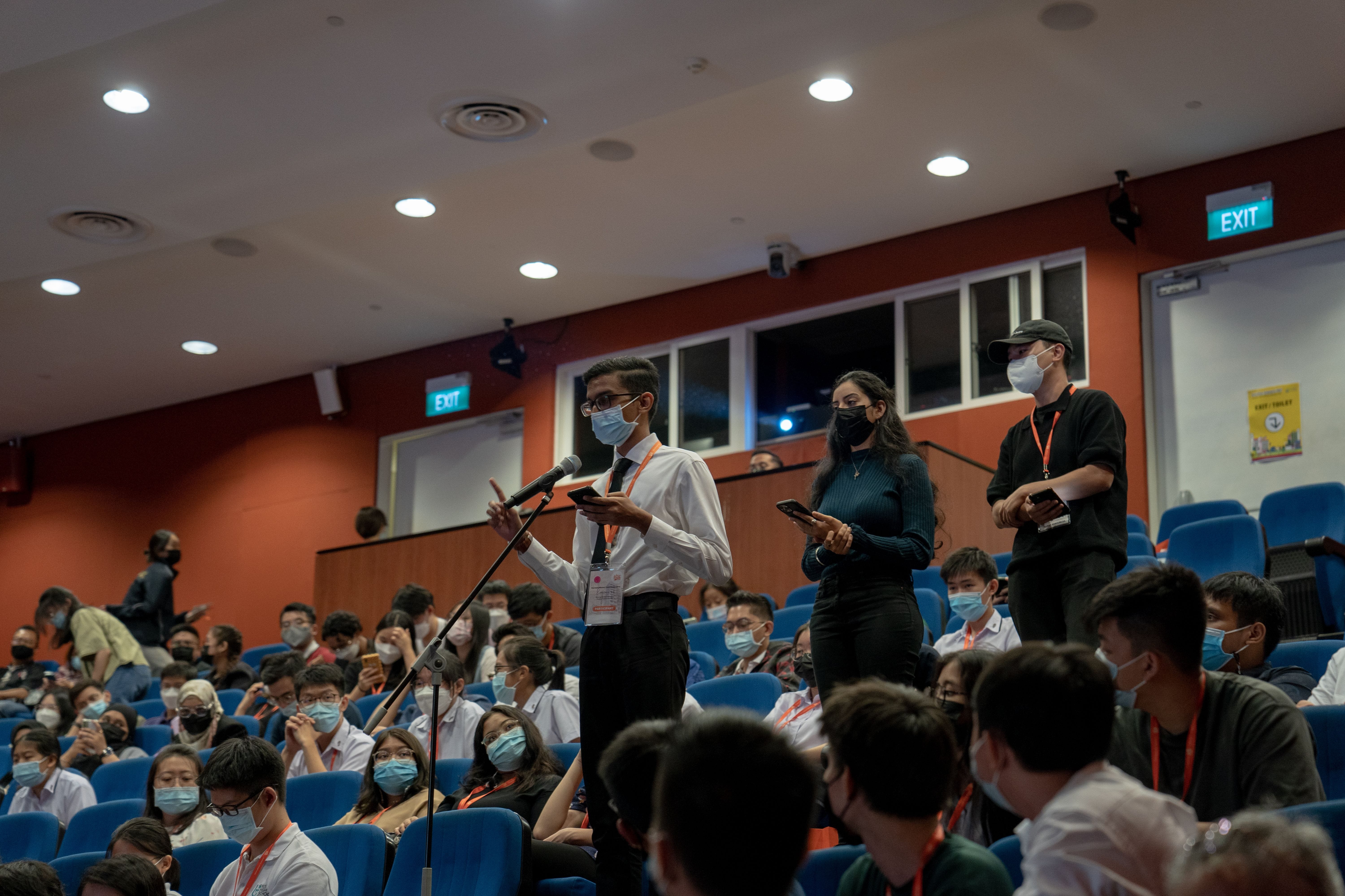 People stand holding notebooks and speaking at a microphone in a crowded auditorium with blue seats.
